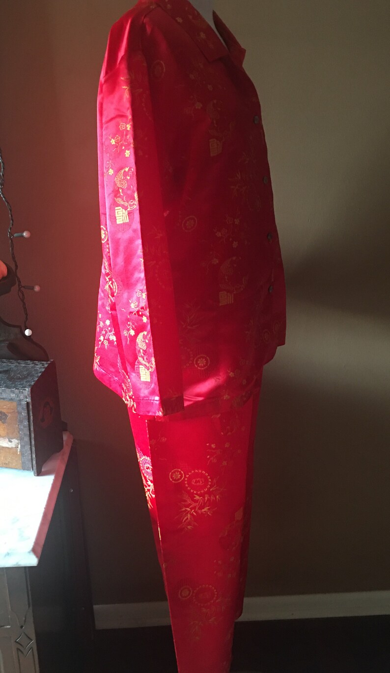 May include: Red pajama set with a button-down shirt and matching pants. The set features a gold floral and character pattern. The fabric has a slight sheen. The pajamas are displayed on a mannequin.