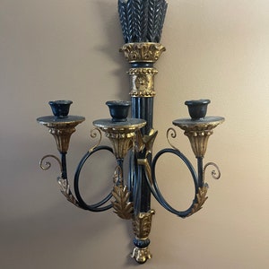 Puede incluir: Un candelero de pared negro y dorado con cuatro brazos para velas. El candelero tiene un diseño de desplazamiento y una parte superior decorativa.