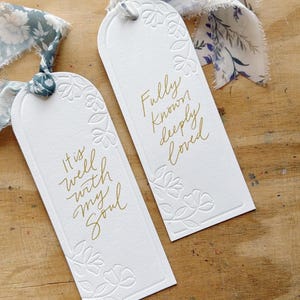 May include: Two white gift tags with gold lettering and floral embossing. One tag reads "It is well with my soul," and the other says "Fully known deeply loved." Each tag has a floral patterned ribbon.