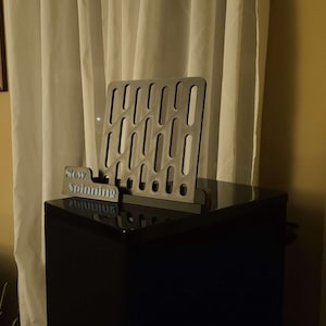May include: A silver-colored vinyl record holder with a rectangular design and oval cutouts. A smaller sign with the words "Stay Spinning" is in front of the holder. The holder sits on top of a black surface.