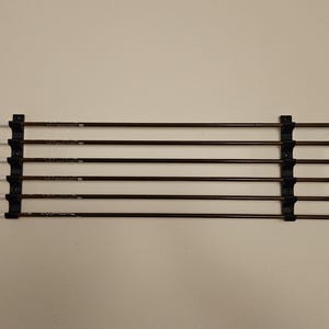 May include: Six arrows with black shafts and white and black fletching are held in a black arrow holder. The arrows are arranged horizontally. The background is a plain, light beige color.