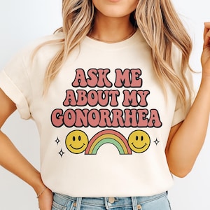May include: Cream-colored t-shirt with the text "ASK ME ABOUT MY GONORRHEA" in pink and brown lettering. The design includes two yellow smiley faces, a rainbow, and four small black stars.