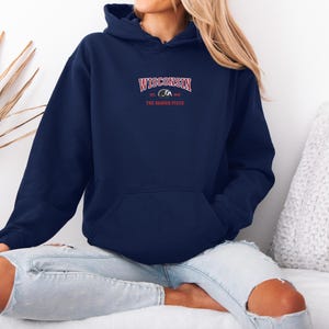 May include: Navy blue hoodie with a front pocket and drawstring hood. The hoodie features the word "WISCONSIN" in red arched text above a badger graphic, with "THE BADGER STATE" below. The sweatshirt is worn by a person.