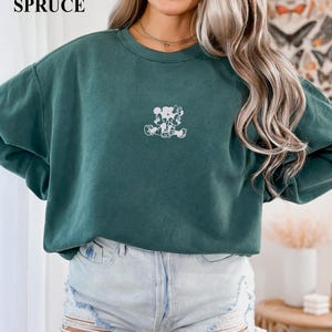May include: A teal-colored sweatshirt with a white graphic of two cartoon characters. The text "BLUE SPRUCE" is in the upper left corner. The sweatshirt is paired with denim shorts.