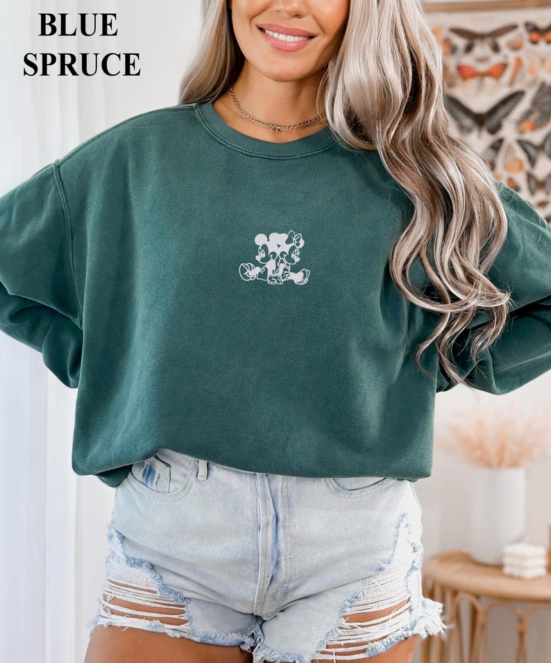 May include: A teal-colored sweatshirt with a white graphic of two cartoon characters. The text "BLUE SPRUCE" is in the upper left corner. The sweatshirt is paired with denim shorts.