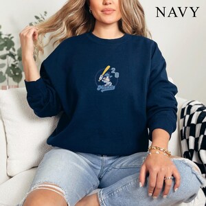 May include: Navy blue sweatshirt featuring an embroidered design of Mickey Mouse holding a baseball bat, with the number 28. The word "Mickey" is written in a script font. The word "NAVY" is printed in white in the upper right corner.