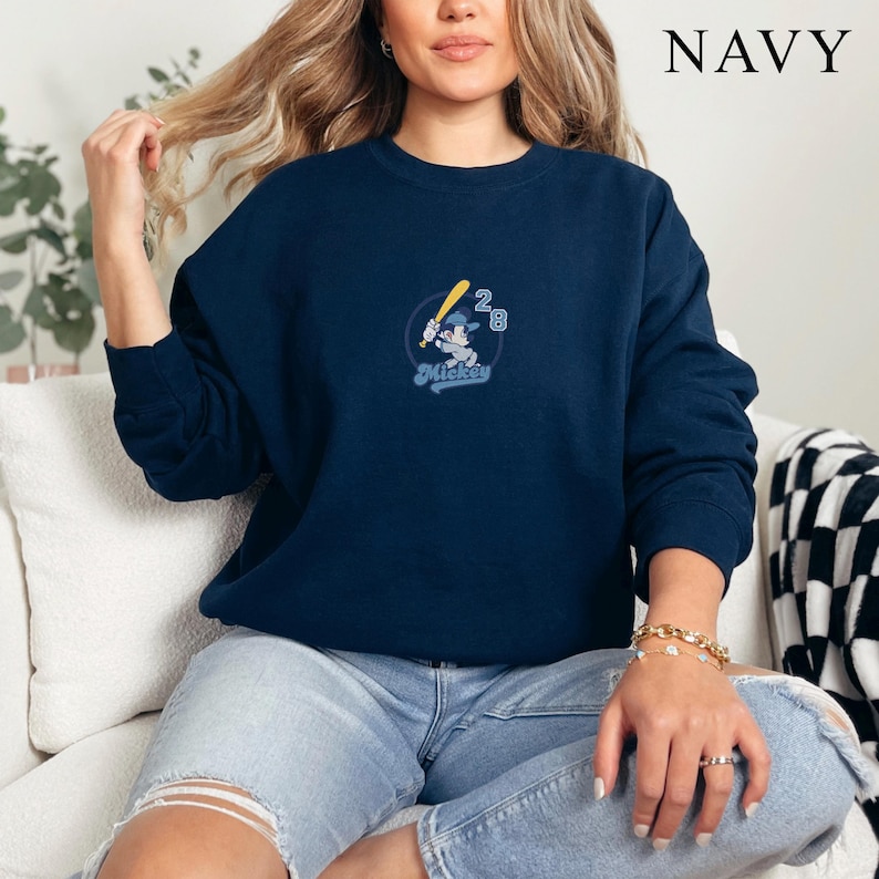 May include: Navy blue sweatshirt featuring an embroidered design of Mickey Mouse holding a baseball bat, with the number 28. The word "Mickey" is written in a script font. The word "NAVY" is printed in white in the upper right corner.