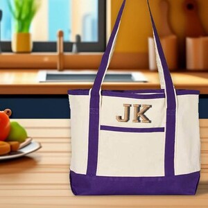 May include: A canvas tote bag with a cream-colored body and purple accents. The bag features a front pocket and long handles. The initials "JK" are embroidered on the front in a gold color. The bag is sitting on a wooden surface.