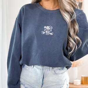 May include: Denim blue sweatshirt with a small, light-colored graphic of Mickey and Minnie Mouse. The sweatshirt has a crew neck and long sleeves. The word "DENIM" is printed in the upper left corner.