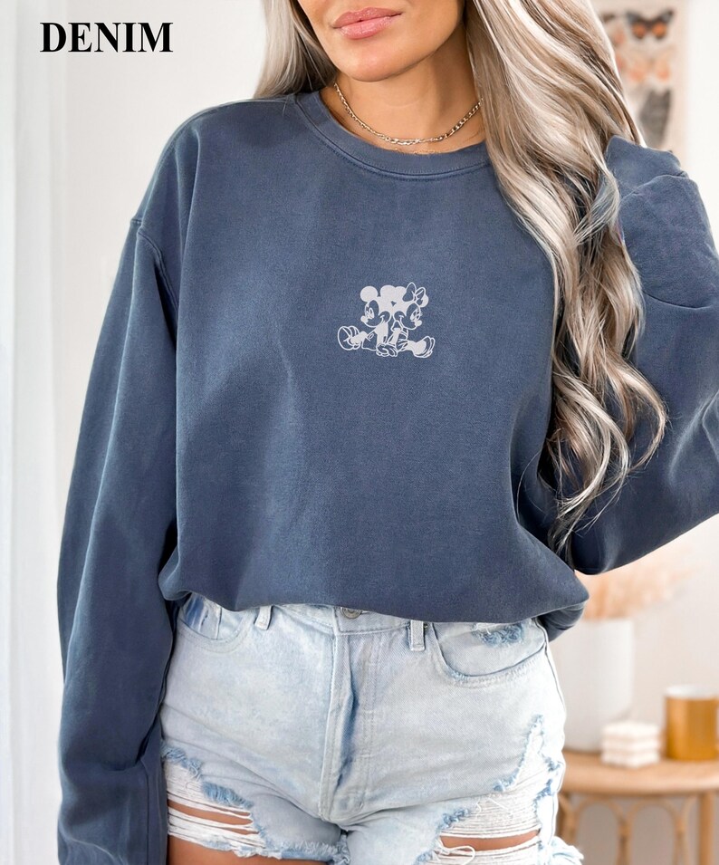 May include: Denim blue sweatshirt with a small, light-colored graphic of Mickey and Minnie Mouse. The sweatshirt has a crew neck and long sleeves. The word "DENIM" is printed in the upper left corner.