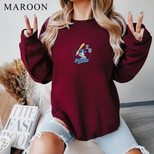 May include: Maroon sweatshirt with a baseball-themed Mickey Mouse graphic. The graphic features Mickey Mouse holding a baseball bat, with the number "28" and the word "Mickey" in a blue font. The word "MAROON" is at the top left.