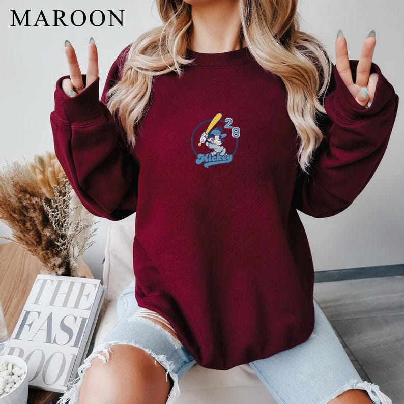May include: Maroon sweatshirt with a baseball-themed Mickey Mouse graphic. The graphic features Mickey Mouse holding a baseball bat, with the number "28" and the word "Mickey" in a blue font. The word "MAROON" is at the top left.