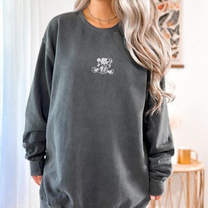 May include: A dark gray sweatshirt with a small, light-colored graphic of a cartoon character on the front. The sweatshirt has long sleeves and a crew neck. The word "PEPPER" is printed in white in the upper left corner.