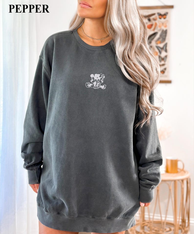 May include: A dark gray sweatshirt with a small, light-colored graphic of a cartoon character on the front. The sweatshirt has long sleeves and a crew neck. The word "PEPPER" is printed in white in the upper left corner.