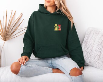 May include: A dark green hoodie with a front pocket and a hood. The hoodie features a small embroidered design of two cartoon cats in yellow and red. The person is wearing light blue jeans.