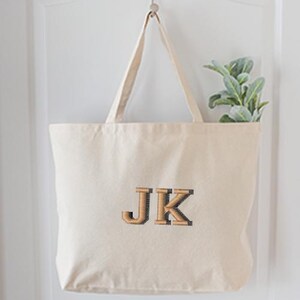 May include: A cream-colored tote bag with the embroidered initials "JK" in a brown, textured font. The bag hangs from a metal hook on a white door. A sprig of green leaves is visible inside the bag.