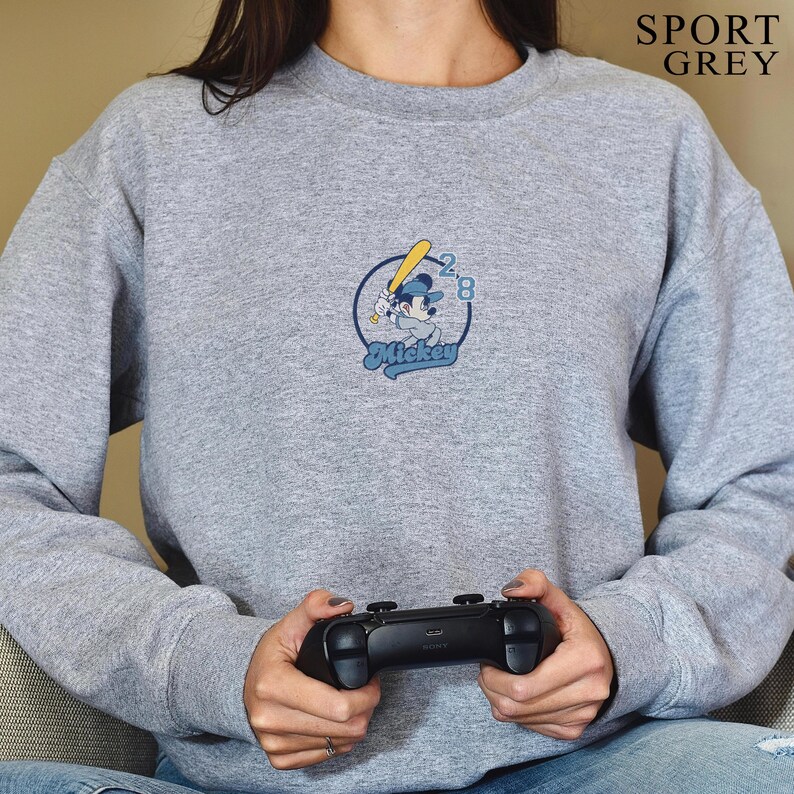 May include: A gray sweatshirt with the text "SPORT GREY" at the top right. The sweatshirt features a circular graphic of Mickey Mouse playing baseball, with the word "Mickey" below. The person is holding a black game controller.
