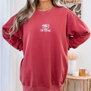 May include: Crimson-colored sweatshirt with a small white graphic of two cartoon characters. The sweatshirt has long sleeves and a crew neck. The word "CRIMSON" is printed in the upper left corner.