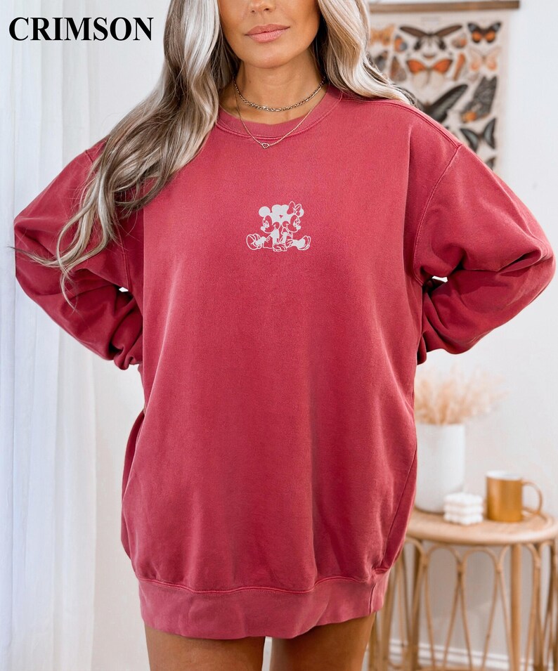 May include: Crimson-colored sweatshirt with a small white graphic of two cartoon characters. The sweatshirt has long sleeves and a crew neck. The word "CRIMSON" is printed in the upper left corner.