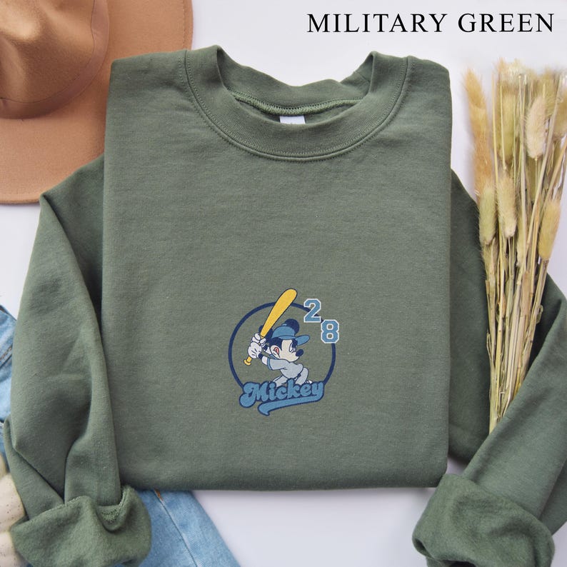 May include: Military green sweatshirt featuring a baseball-themed Mickey Mouse graphic. The design includes Mickey holding a bat, a baseball, and the number 28. The word "Mickey" is written in a script font below the character.
