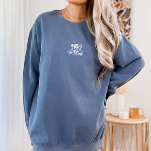 May include: Blue jean colored sweatshirt with a small white graphic on the chest. The sweatshirt has a crew neck and long sleeves. The text "BLUE JEAN" is at the top of the image.