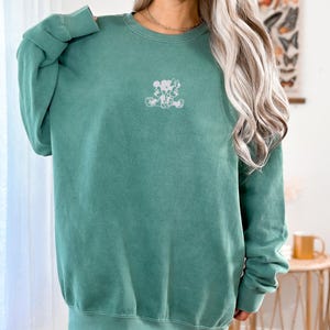 May include: Light green sweatshirt with a crew neck and long sleeves. The sweatshirt features a small, white graphic of cartoon characters on the front. The text "LIGHT GREEN" is at the top of the image.