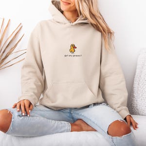May include: A beige hoodie with a kangaroo pocket and drawstring hood. The hoodie features a small embroidered yellow duck with a red beak and the text "got any grapes?" below it. The model is wearing light blue jeans.