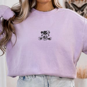 May include: A light purple sweatshirt with a small black graphic of Mickey and Minnie Mouse. The word "ORCHID" is printed in white at the top left. The sweatshirt is paired with light blue denim shorts.