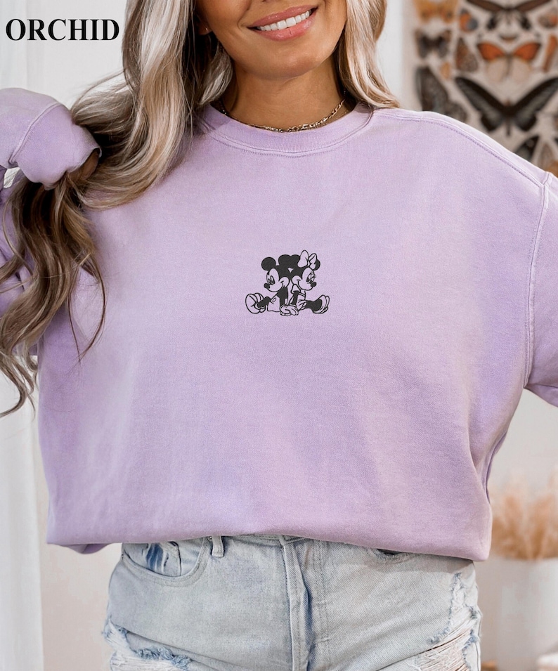 May include: A light purple sweatshirt with a small black graphic of Mickey and Minnie Mouse. The word "ORCHID" is printed in white at the top left. The sweatshirt is paired with light blue denim shorts.