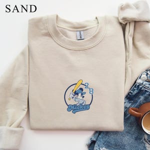 May include: Sand-colored sweatshirt with a crew neck. The front features a circular graphic of Mickey Mouse in a baseball uniform, holding a bat. The graphic includes the text "Mickey" and the numbers "28" in blue.