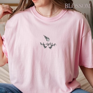 May include: A light pink t-shirt featuring a graphic of dinosaurs, cacti, and a Wi-Fi signal with a shooting star. The word "BLOSSOM" is printed in the upper right corner. The shirt has a crew neck and short sleeves.