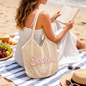 Puede incluir: Un bolso tote de crochet beige con el nombre "Sophia" bordado en rosa. La bolsa está sobre una toalla de playa a rayas azules y blancas. Un sombrero de paja y gafas de sol están cerca, sugiriendo un día de playa.