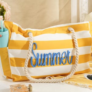 以下が含まれることがあります： 黄色と白のストライプのトートバッグで、青色で「Summer」の文字が刺繍されています。バッグにはロープのハンドルとサイドポケットが付いています。ライトブルーのタンブラーがサイドポケットに入っています。バッグは白い表面にあります。