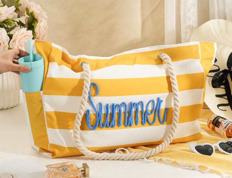 以下が含まれることがあります： 黄色と白のストライプのトートバッグで、青色で「Summer」の文字が刺繍されています。バッグにはロープのハンドルとサイドポケットが付いています。ライトブルーのタンブラーがサイドポケットに入っています。バッグは白い表面にあります。