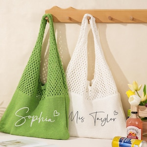 May include: Two crochet tote bags, one green and one white, hang from a wooden rack. The green bag has the name "Sophia" in script, and the white bag says "Miss Taylor". Both bags have long handles.