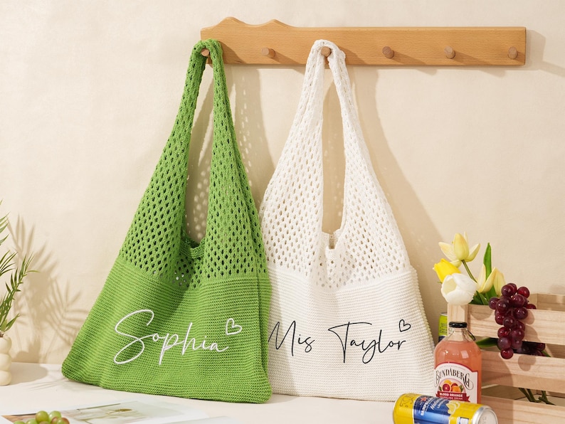May include: Two crochet tote bags, one green and one white, hang from a wooden rack. The green bag has the name "Sophia" in script, and the white bag says "Miss Taylor". Both bags have long handles.