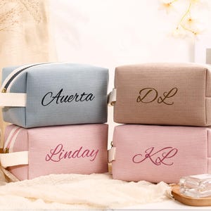 Puede incluir: Cuatro neceseres personalizados en colores pastel: azul claro, beige y dos tonos de rosa. Cada neceser tiene cierre de cremallera, asa e iniciales o nombre bordados. Los neceseres están apilados.