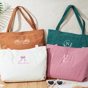 May include: Four corduroy tote bags in shades of brown, white, teal, and pink. Each bag has long handles and embroidered details, including names, initials, and floral designs. The bags are displayed on a wooden surface.