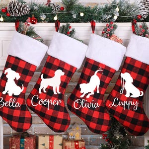May include: Four Christmas stockings with a red and black buffalo plaid pattern. Each stocking has a white silhouette of a dog and a name: Bella, Cooper, Olivia, and Luppy. The stockings have white faux fur cuffs.