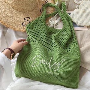 May include: A green knitted tote bag with the name "Emily" embroidered in white script. The date "09-12-2023" is below the name. The bag has a large open weave design and a handle. A straw hat and a beaded purse are in the background.