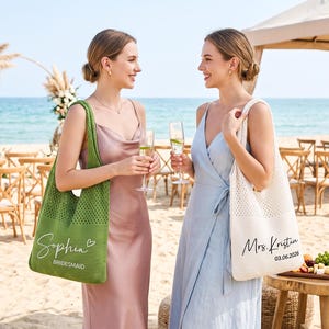 May include: Two women at a beach wedding, each holding a champagne flute. One carries a green tote bag with "Sophia Bridesmaid" printed on it. The other carries a white tote bag with "Mrs. Kristin 03.06.2026" printed on it. Both are wearing dresses.