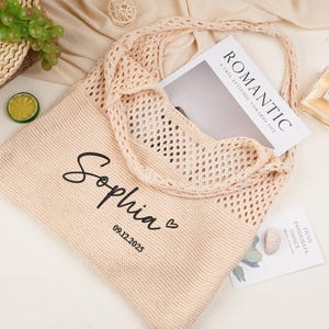 May include: A light peach knitted tote bag with a mesh overlay. The bag is personalised with the name "Sophia" and the date "09.12.2025". A magazine with the word "ROMANTIC" is inside the bag. A bottle of perfume and some fruit are also visible.