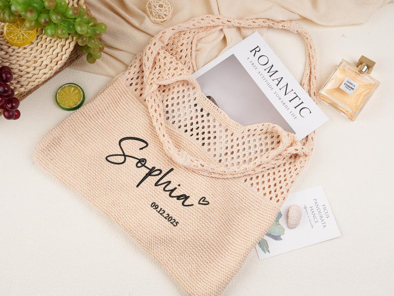 May include: A light peach knitted tote bag with a mesh overlay. The bag is personalised with the name "Sophia" and the date "09.12.2025". A magazine with the word "ROMANTIC" is inside the bag. A bottle of perfume and some fruit are also visible.