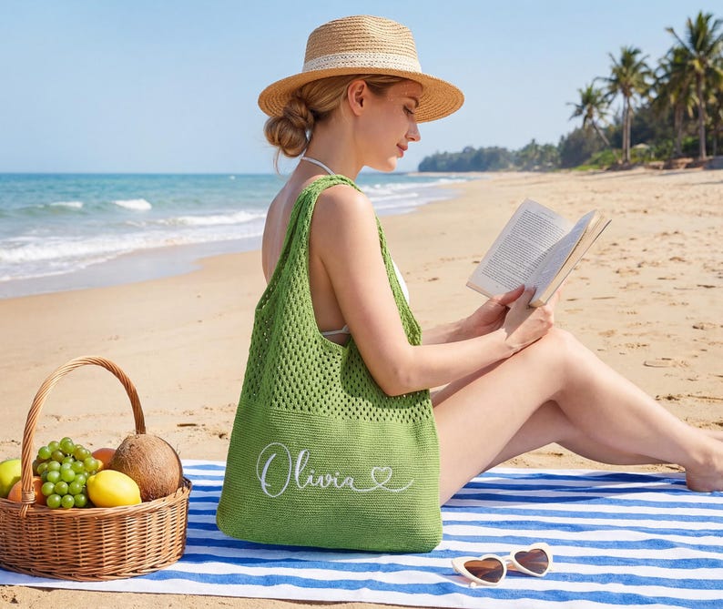 May include: A green, open-weave tote bag with the name "Olivia" embroidered in white, on a blue and white striped beach towel. A straw hat and heart-shaped sunglasses are also visible. A wicker basket filled with fruit sits nearby.
