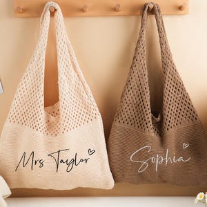 May include: Two knitted tote bags, one cream and one brown, hang against a light beige wall. The cream bag has "Mrs Taylor" written on it, and the brown bag has "Sophia" written on it. Both bags have a heart symbol next to the name.