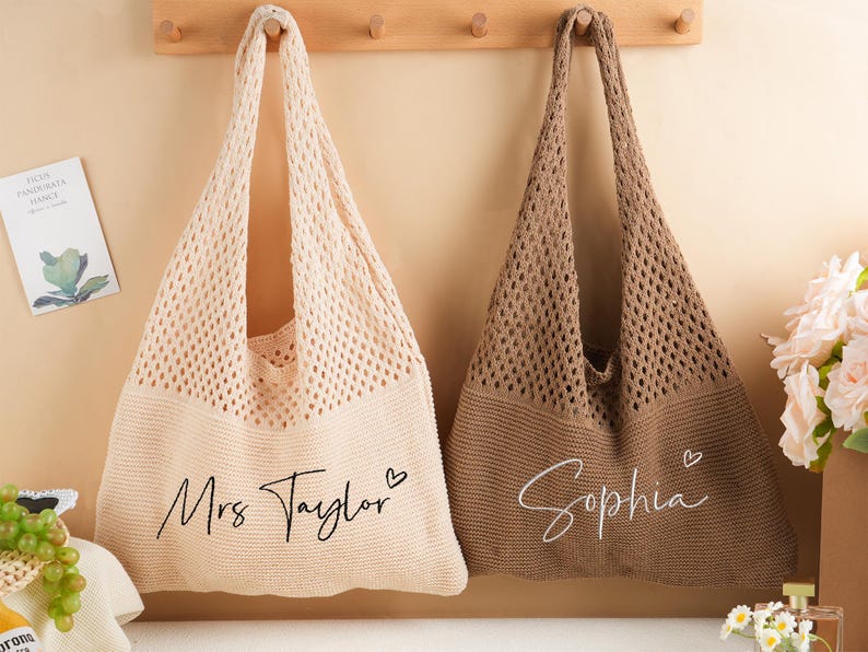 May include: Two knitted tote bags, one cream and one brown, hang against a light beige wall. The cream bag has "Mrs Taylor" written on it, and the brown bag has "Sophia" written on it. Both bags have a heart symbol next to the name.