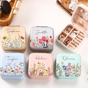 Może przedstawiać: Kolekcja spersonalizowanych pudełek na biżuterię w różnych pastelowych kolorach, każde ozdobione kwiatowymi wzorami i imieniem. Jedno pudełko jest otwarte, ukazując przegródki na pierścionki, kolczyki i naszyjniki. Pudełka są kwadratowe z zapięciem na zamek.