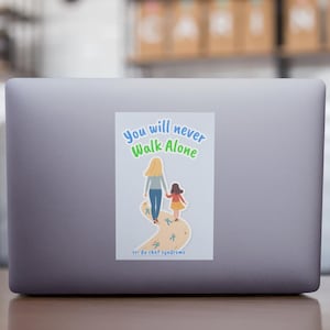 May include: A sticker on a laptop features the text "You will never Walk Alone" above an illustration of a woman and child walking together. The sticker has a light blue background with a path and green accents.