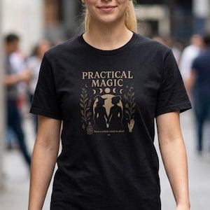 Puede incluir: Una camiseta negra de manga corta con un estampado gráfico dorado. El diseño incluye el texto "PRACTICAL MAGIC" sobre dos siluetas de figuras tomadas de la mano, rodeadas de elementos botánicos, fases lunares y un icono de mano.