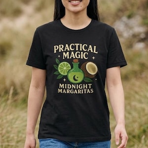 May include: Black t-shirt with the words "PRACTICAL MAGIC" above an illustration of a lime, a potion bottle, and a coconut. Below the illustration is the text "MIDNIGHT MARGARITAS". The person is wearing blue jeans.